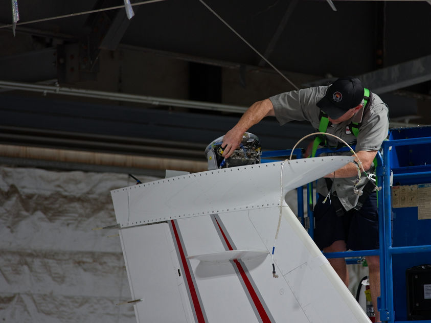 A technician on a hydraulic lift performs maintenance on the tail section of an aircraft inside a hangar with visible structural elements.