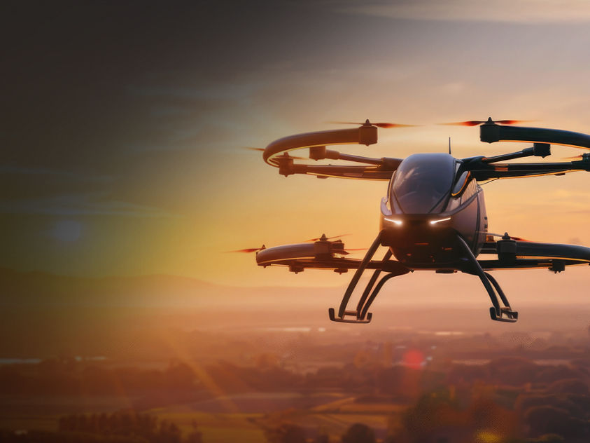 A multi-rotor drone captured mid-flight during sunset, with warm lighting and a scenic backdrop of fields, trees, and hills.