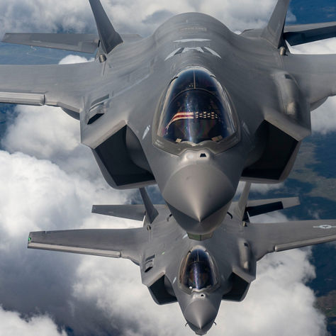 A pair of grey military jets soar in formation above the clouds, with Earth’s surface faintly visible below.