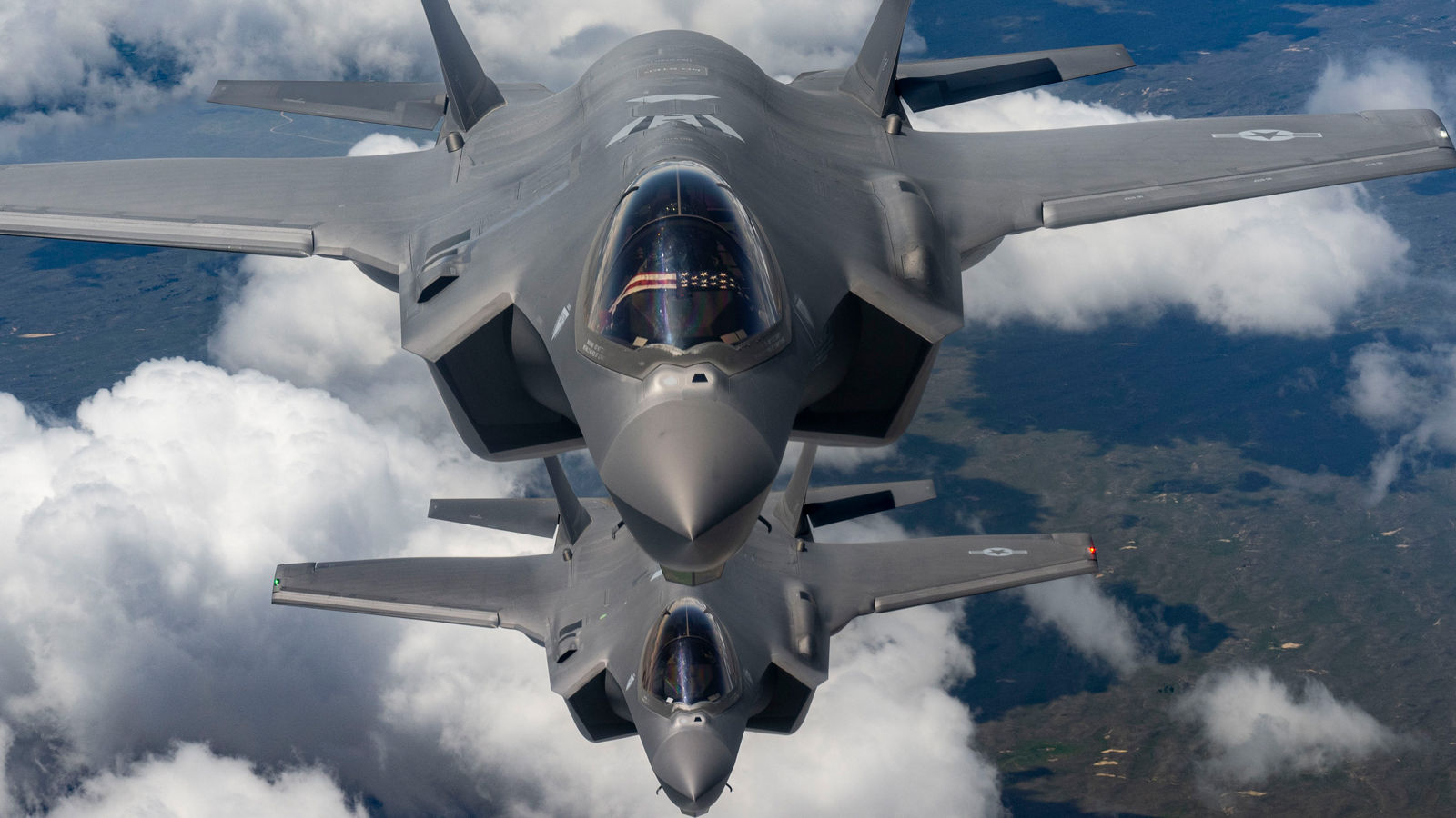 A pair of grey military jets soar in formation above the clouds, with Earth’s surface faintly visible below.