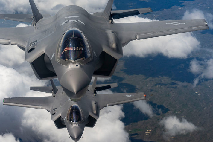 A pair of grey military jets soar in formation above the clouds, with Earth’s surface faintly visible below.