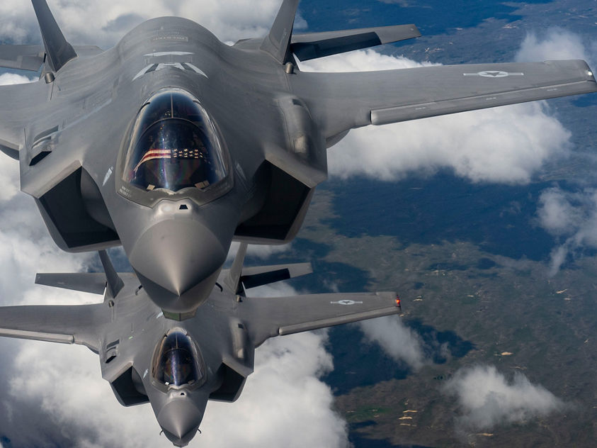 A pair of grey military jets soar in formation above the clouds, with Earth’s surface faintly visible below.