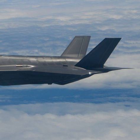 A sleek gray military aircraft with twin tail fins soars above a cloud layer, with a pilot visible in the cockpit.
