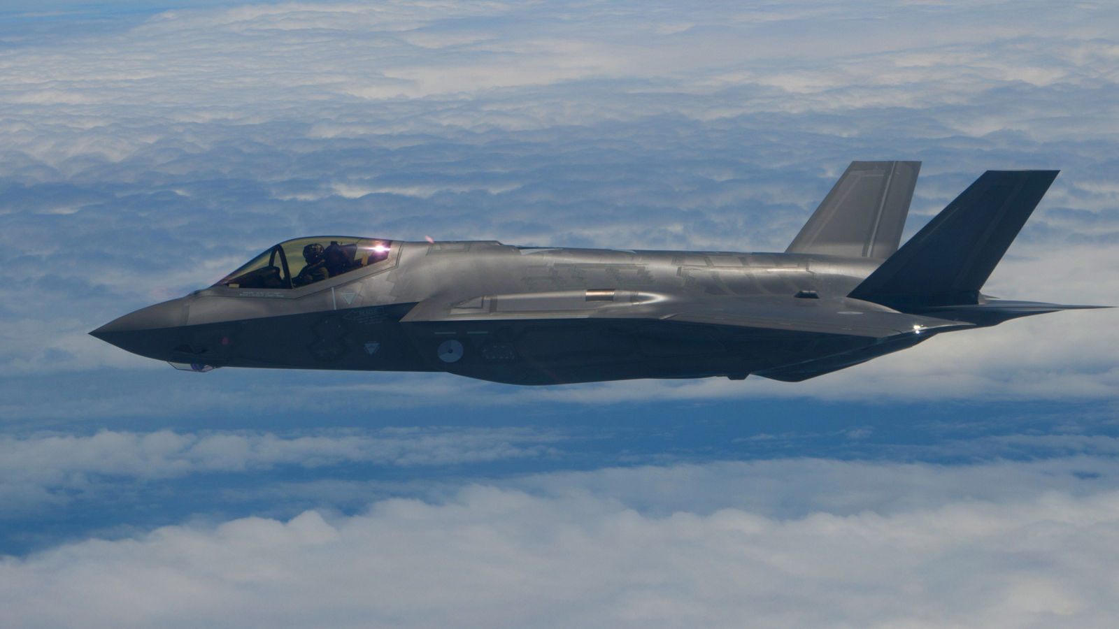A sleek gray military aircraft with twin tail fins soars above a cloud layer, with a pilot visible in the cockpit.