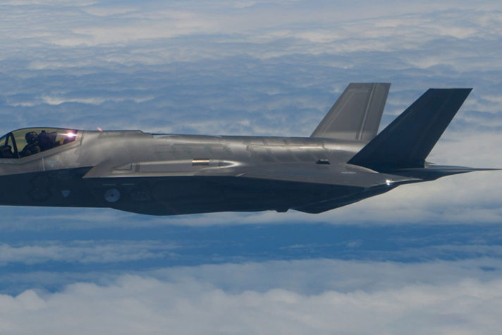 A sleek gray military aircraft with twin tail fins soars above a cloud layer, with a pilot visible in the cockpit.
