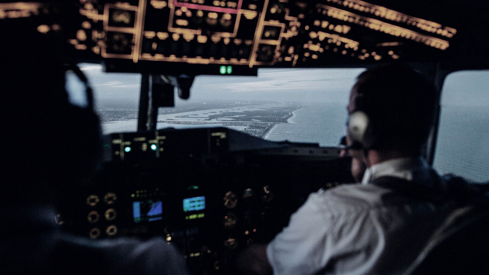 A cockpit view of two pilots guiding an aircraft along the coastline during approach, with flight instruments illuminated and a detailed shoreline visible through the windshield. Ideal for aviation safety, navigation technology, and flight operations content.