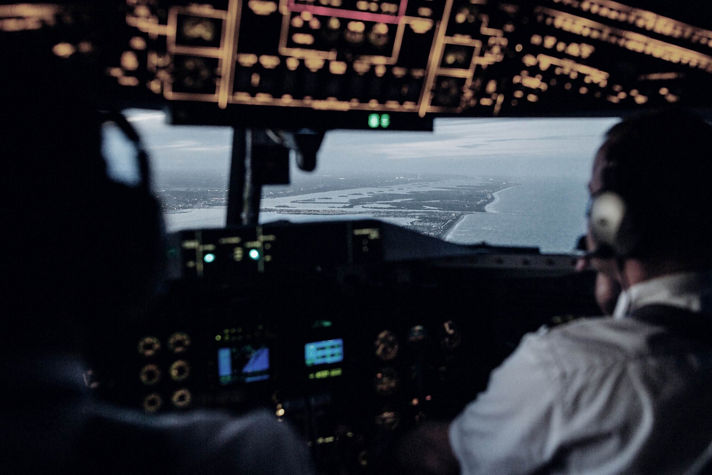 A cockpit view of two pilots guiding an aircraft along the coastline during approach, with flight instruments illuminated and a detailed shoreline visible through the windshield. Ideal for aviation safety, navigation technology, and flight operations content.