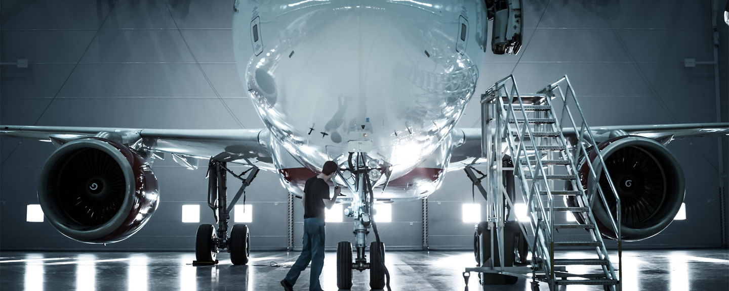 Front view of a commercial aircraft inside a hangar while a technician performs inspection near the landing gear with access equipment nearby
