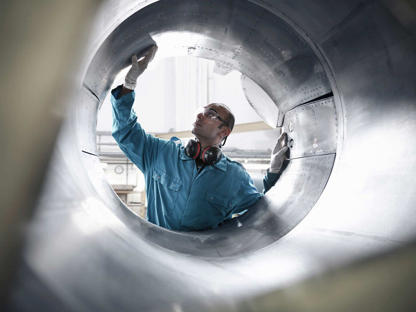 A male aerospace technician in a blue coverall with hearing protection inspects the interior of a large cylindrical aircraft engine nacelle from inside, viewed through the opening. This image represents Honeywell's engine maintenance and inspection services.