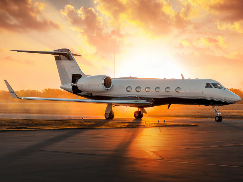 The image shows a large-cabin private business jet, consistent with a Gulfstream G450 or G550, parked on a runway at sunset. 