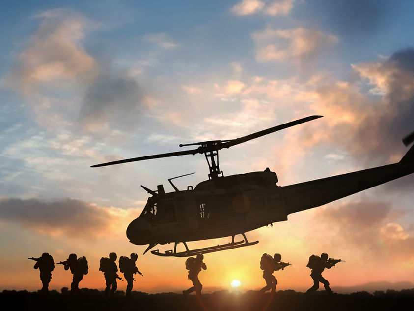 Silhouette of a military helicopter flying above soldiers walking in formation during sunset with orange and blue hues.