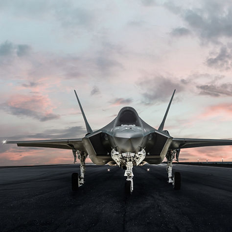 Front view of a modern stealth fighter jet parked on a runway at dusk beneath a pink and gray cloud-filled sky.