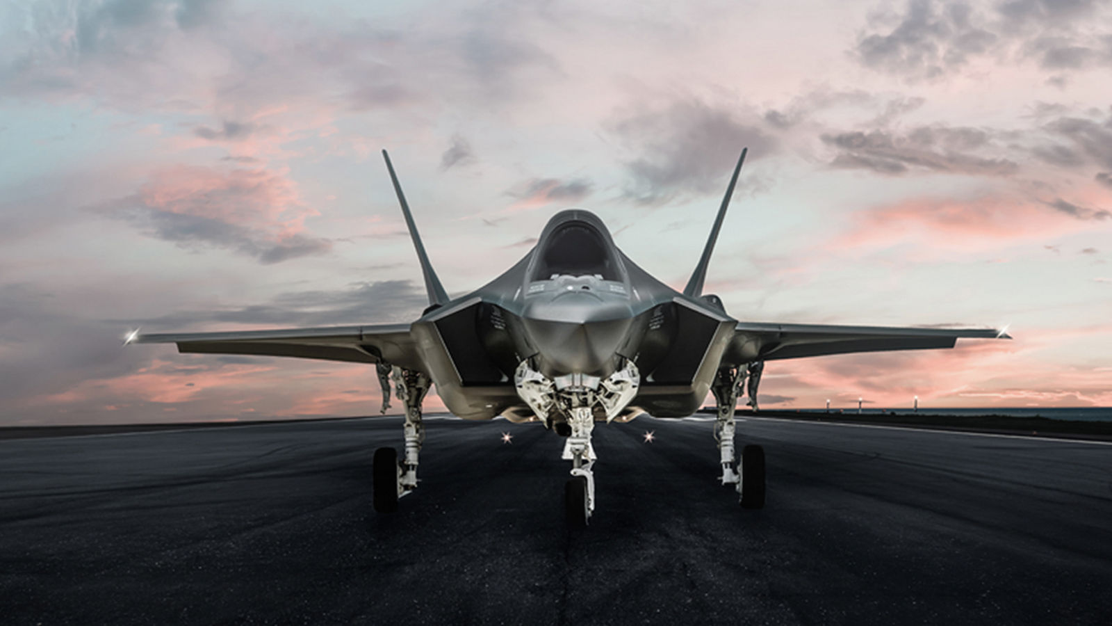 Front view of a modern stealth fighter jet parked on a runway at dusk beneath a pink and gray cloud-filled sky.