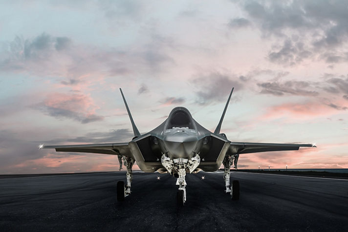 Front view of a modern stealth fighter jet parked on a runway at dusk beneath a pink and gray cloud-filled sky.