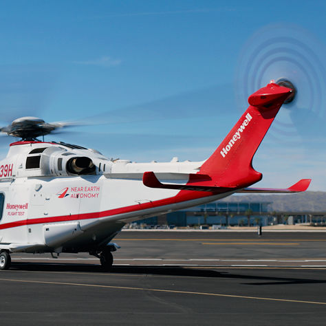 White and red helicopter with rotors spinning, branded with Honeywell and Near Earth Autonomy, parked near an airport control tower.