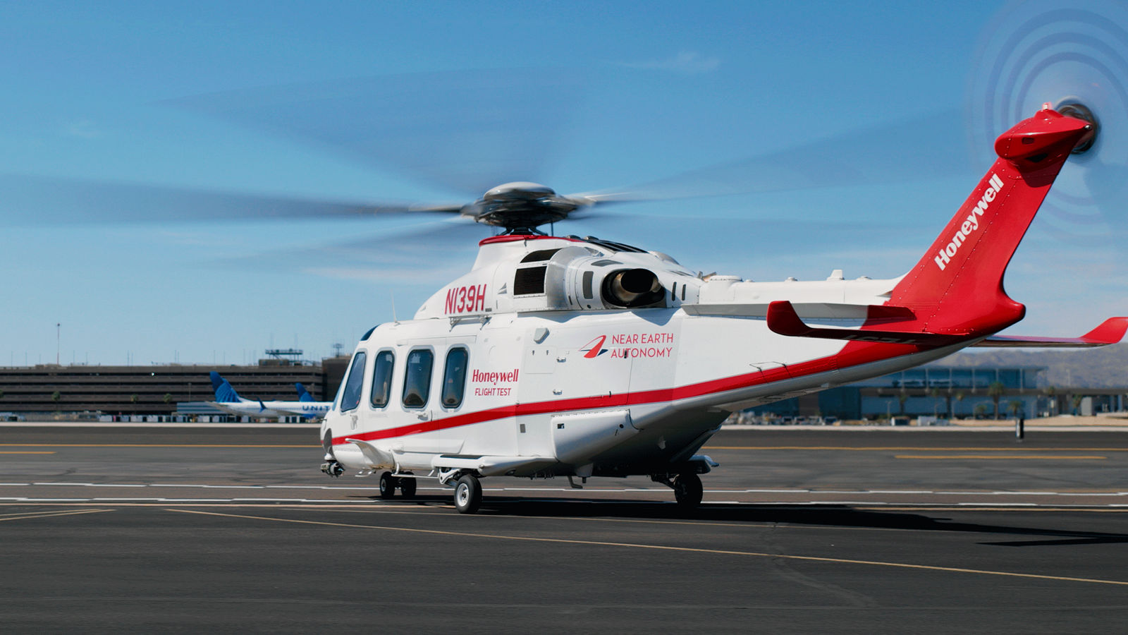 White and red helicopter with rotors spinning, branded with Honeywell and Near Earth Autonomy, parked near an airport control tower.