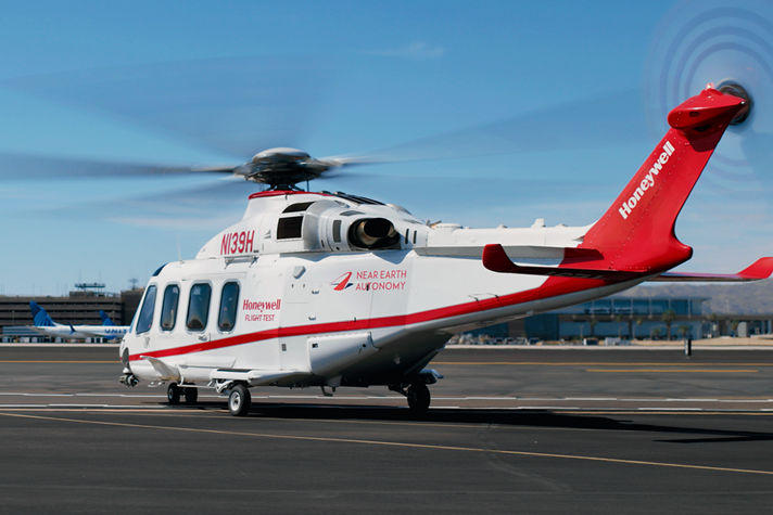 White and red helicopter with rotors spinning, branded with Honeywell and Near Earth Autonomy, parked near an airport control tower.