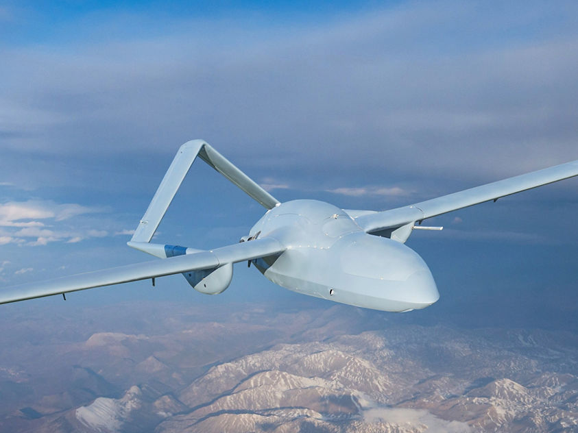an image of an U.S. military drone flies over rugged mountain terrain surveying the landscape below.