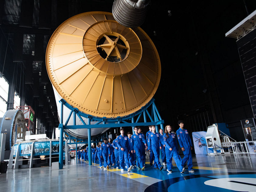 A group of students in blue flight suits walk through a large aerospace exhibit featuring a massive rocket component. The image captures exploration, STEM education, and space‑focused learning.
