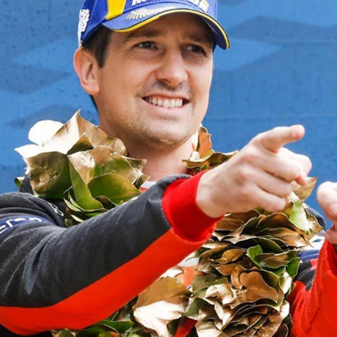 A person in a racing suit and laurel wreath celebrates with both hands pointing forward, set against a blue patterned background.
