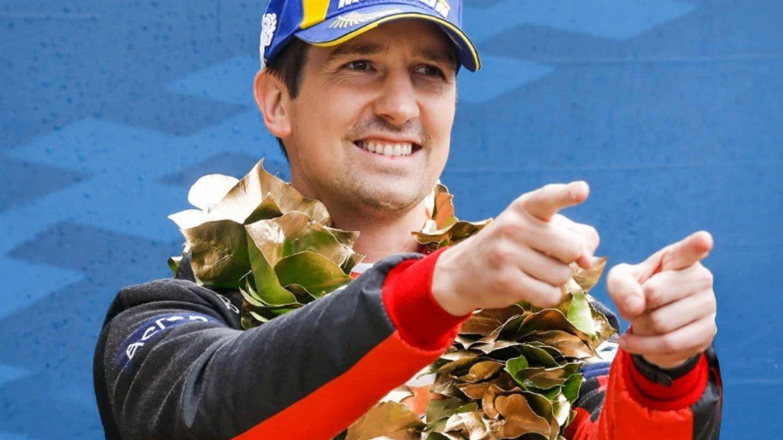 A person in a racing suit and laurel wreath celebrates with both hands pointing forward, set against a blue patterned background.