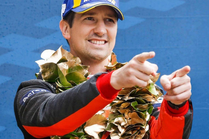 A person in a racing suit and laurel wreath celebrates with both hands pointing forward, set against a blue patterned background.