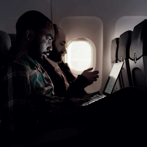 Airline passengers seated in a dim cabin working on a laptop and mobile device during flight.