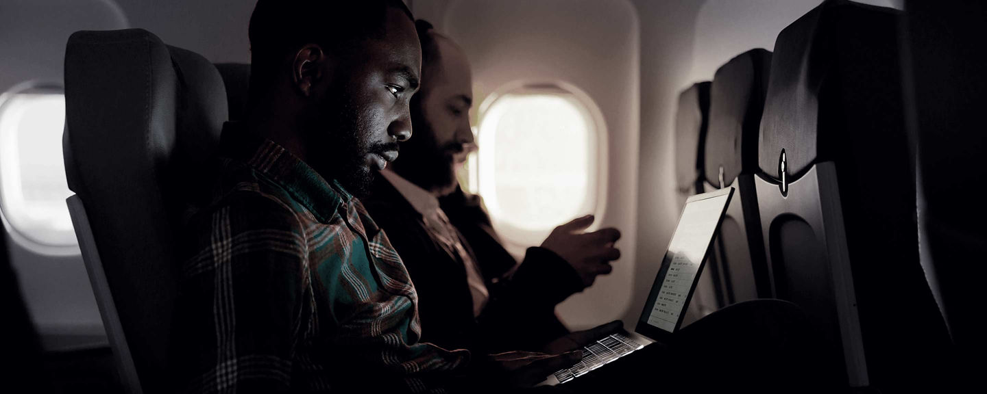 Airline passengers seated in a dim cabin working on a laptop and mobile device during flight.