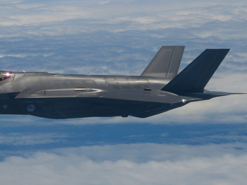 A sleek gray military aircraft with twin tail fins soars above a cloud layer, with a pilot visible in the cockpit.