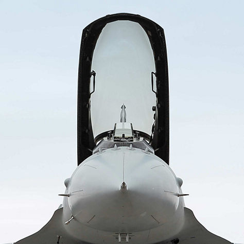Dramatic upward-looking front view of a fighter jet with the cockpit canopy fully open, showing the ejection seat and cockpit interior, captured against a white sky. This defense image represents Honeywell's fighter jet avionics, mission systems, and cockpit integration.