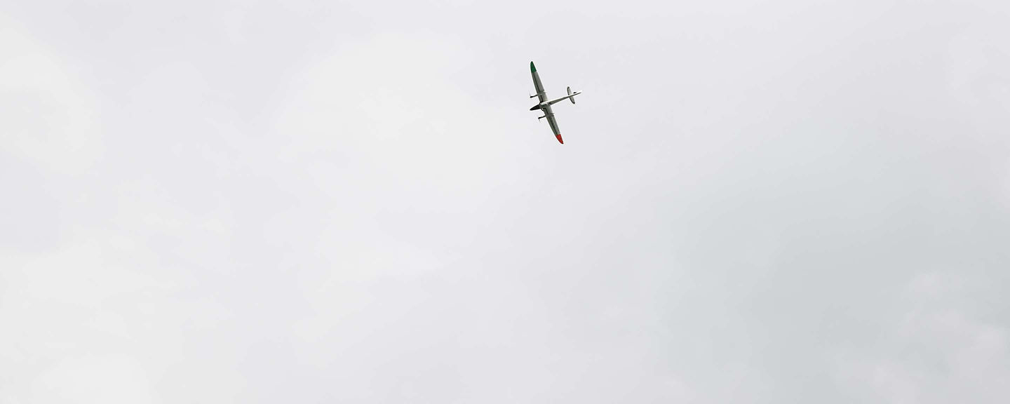 A small glider or UAV is captured as a tiny silhouette flying in a wide overcast grey sky, emphasized by the vast empty sky around it. The minimalist composition suggests Honeywell's unmanned systems and UAV navigation technology in low-signature environments.