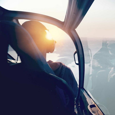 Pilot seated in a helicopter cockpit with warm sunlight illuminating the canopy and distant landscape.