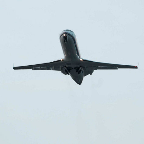 Upward view of a dark business jet climbing away from the camera against an overcast white sky, showing the underside of the fuselage, tail section, and engine pods as the aircraft departs. This image reflects Honeywell's business jet avionics and performance solutions.