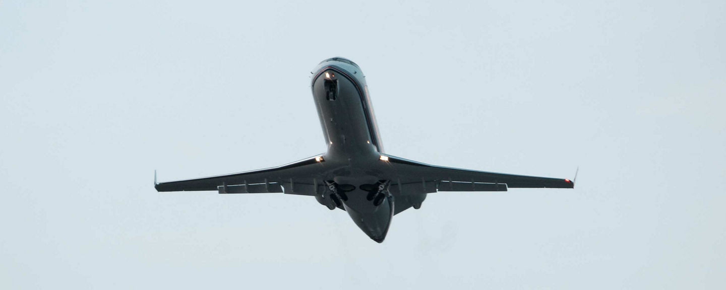 Upward view of a dark business jet climbing away from the camera against an overcast white sky, showing the underside of the fuselage, tail section, and engine pods as the aircraft departs. This image reflects Honeywell's business jet avionics and performance solutions.