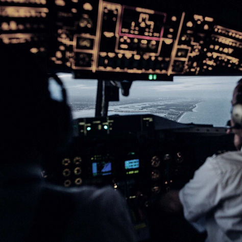 Rear cockpit view of pilots managing instruments while flying along a coastal route.