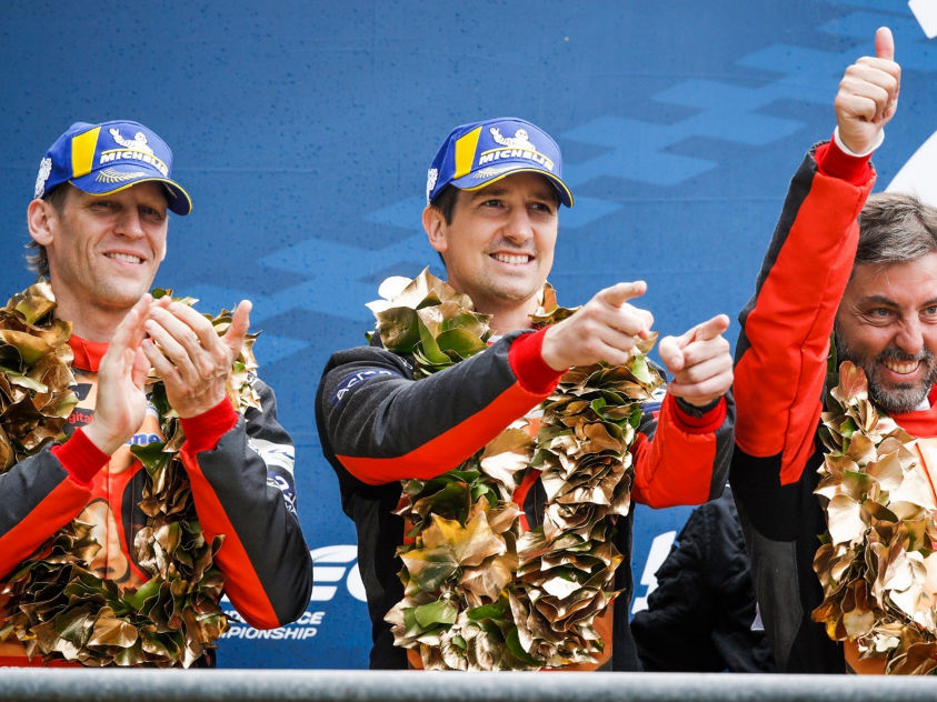 Three racers in suits and caps celebrate on a podium with wreaths, thumbs-up and pointing gestures, set against a championship backdrop.